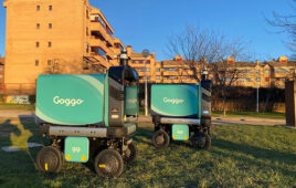 Two blue, boxy mobile robots rolling across a lawn with buildings in the background.