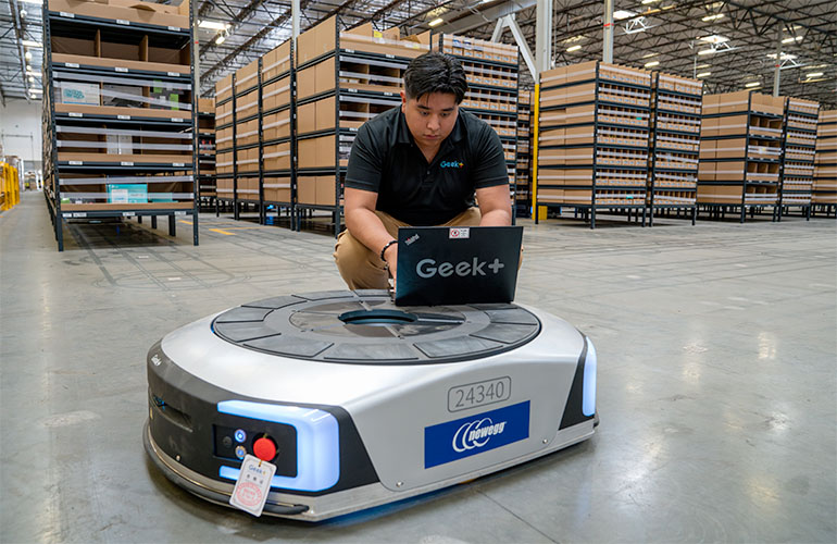 an engineer updates software on a Geek+ mobile robot with a laptop in the middle of a warehouse.